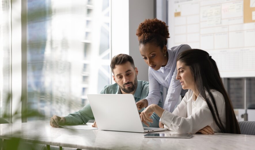 Drei Personen arbeiten gemeinsam konzentriert an einem Laptop und besprechen etwas im modernen Büro.